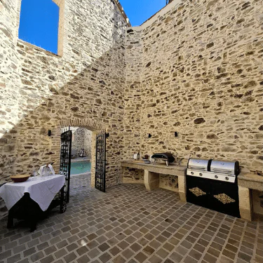 Outdoor kitchen with a brick floor, stone walls, a grill, worktop, and a table set with crockery. Open iron gate leads to a pool area outside under a clear blue sky.