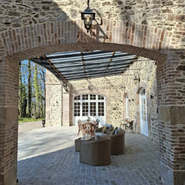 Stone patio with archways, wicker chairs, and a dining table under a glass roof. The area is surrounded by stone walls, lantern lights, and trees visible in the background.