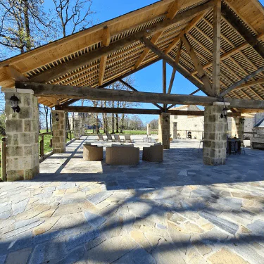 Open wooden pavilion with a pitched roof, stone pillars, and outdoor seating, set on a stone patio. Trees and stone buildings are visible in the sunny background.
