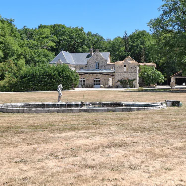 A stone house sits behind a dry, empty fountain featuring a statue in the centre, surrounded by brown grass and trees under a clear blue sky.