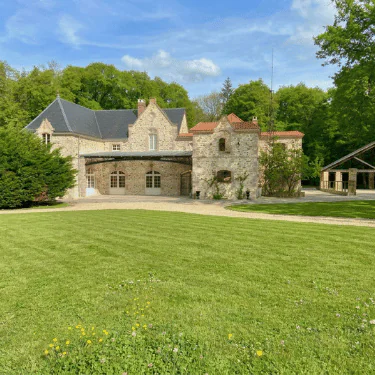 A stone mansion with arched windows and a red-tiled roof stands on a well-manicured lawn, surrounded by green trees under a blue sky with scattered clouds.