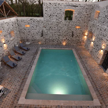 Rectangular swimming pool surrounded by stone walls, five sun loungers, and wall-mounted lights, creating a cosy outdoor courtyard atmosphere in the evening.
