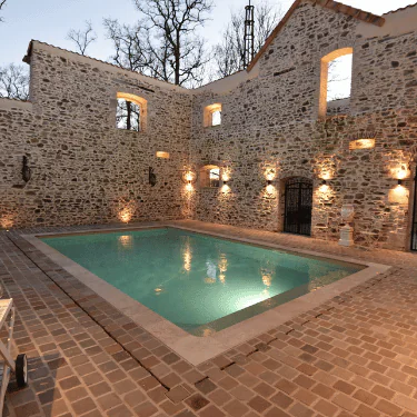 A rectangular indoor pool is surrounded by brick walls with windows and soft yellow lights, creating a cosy and rustic atmosphere. The setting appears to be part of a restored stone building.