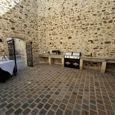 Outdoor kitchen with stone walls, built-in worktops, a grill, and a pizza oven. An iron gate opens to a pool area, and there's a table on the left with a white tablecloth. The floor is made of brown stone tiles.
