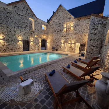 A stone courtyard with an illuminated pool, loungers, rolled towels, and a small table with a wine bottle and glasses at dusk.