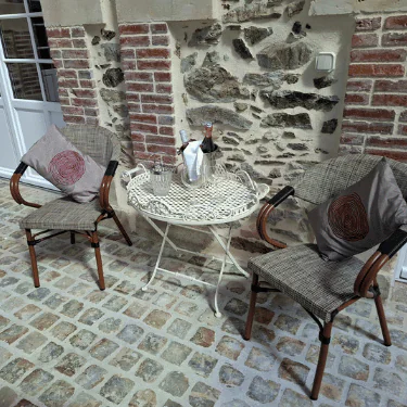 Two cushioned chairs and a round metal table with a wine bottle in an ice bucket, two glasses, and a jug are set on a cobblestone patio against a brick and stone wall.