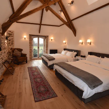 A spacious bedroom with two large beds, exposed wooden beams, a stone wall, wooden floor, a red rug, an armchair, and double doors leading outside.