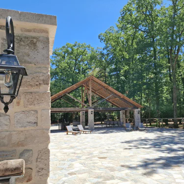 A stone patio with a lantern on a wall in the foreground and a wooden open-air pavilion surrounded by trees in the background on a sunny day.