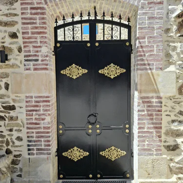 A large, ornate black metal door with gold decorative accents is set in a stone and brick wall. The door has a ring handle and decorative ironwork on the arched fanlight above.