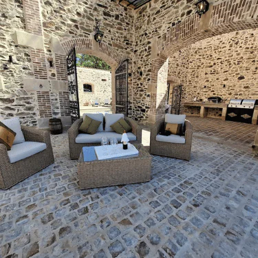 Wicker garden furniture with cushions and a coffee table sit on a cobblestone floor in a rustic stone courtyard. Open gates and a barbecue area are visible in the background.