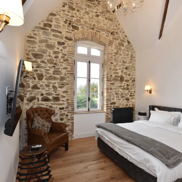 A cosy bedroom with a stone feature wall, large window, brown leather armchair, wooden floor, double bed with white bed linen, and modern and classic light fittings.