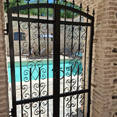Decorative wrought iron gate with scrollwork and latch, set in a stone wall, opens to reveal a clear blue swimming pool and patio area under a sunny sky.