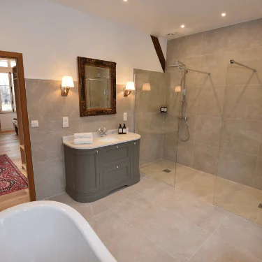 Modern bathroom with large beige tiles, a glass walk-in shower, a grey vanity with a mirror, two wall lights, and part of a white bath in the foreground.