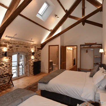 Cosy bedroom with two beds, rustic exposed wooden beams, stone feature wall, skylight, open wardrobe, and warm lighting, creating an inviting and comfortable atmosphere.