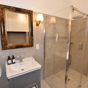 A modern bathroom with a glass shower, grey vanity, white basin, framed mirror, wall light, and two brown soap bottles on the basin. The walls and floor are tiled in light grey.