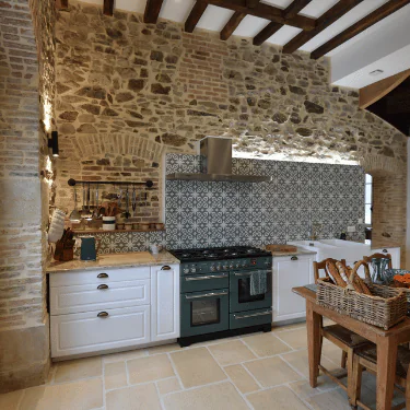 A rustic kitchen features stone walls, patterned tile splashback, a green cooker, white cupboards, and a wooden table with a wicker basket. Wooden beams span the ceiling, adding to the cosy, country ambience.