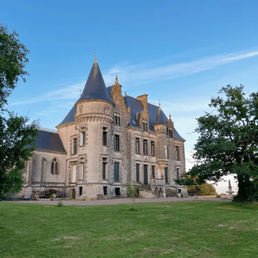A grand stone château with pointed towers stands on a grassy lawn, surrounded by trees under a clear blue sky.