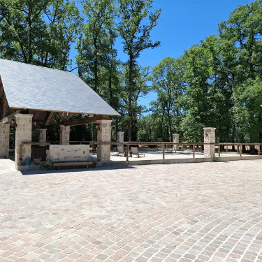 Open paved area with a small stone shelter and picnic tables, surrounded by a wooden fence and tall trees under a clear blue sky.