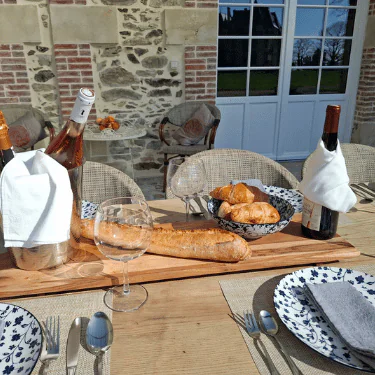 A sunlit outdoor table set for a meal with wine, baguettes, croissants, glasses, plates, and cutlery. Stone wall and glass doors in the background.
