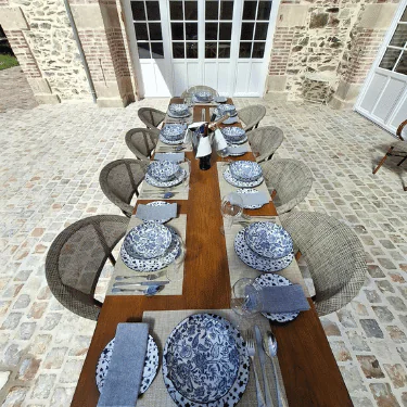 Outdoor dining table set for ten with blue and white patterned plates, glasses, cutlery, and grey napkins on placemats, surrounded by wicker chairs on a stone patio.
