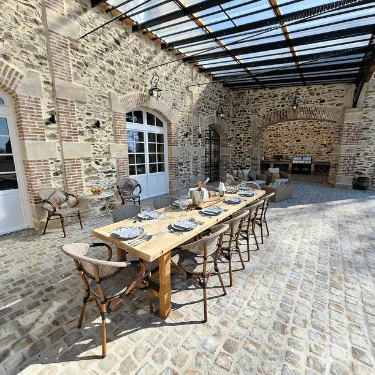 A rustic outdoor dining area with a wooden table set for eight, surrounded by wicker chairs, under a glass roof in a stone courtyard.
