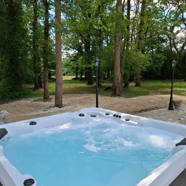 A bubbling hot tub sits outdoors on a stone patio, surrounded by tall green trees and grassy areas, with lamp posts nearby under a clear sky.