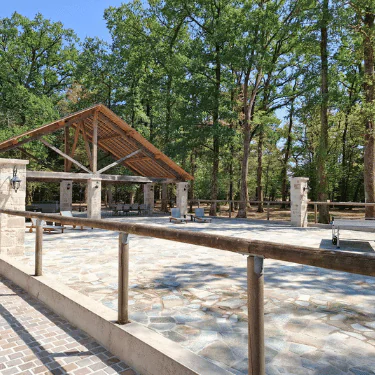 A stone-paved outdoor area with a wooden-roofed pavilion sits among tall trees, surrounded by a wooden railing and stone pillars on a sunny day.