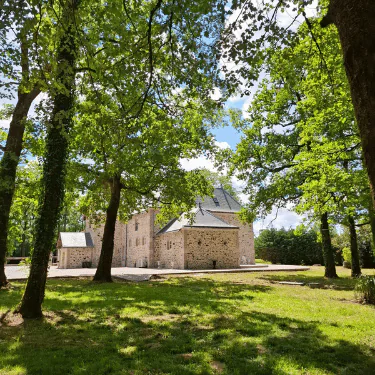 A stone building with a sloping roof stands among tall, leafy trees on a sunny day, casting shadows on the green grass.
