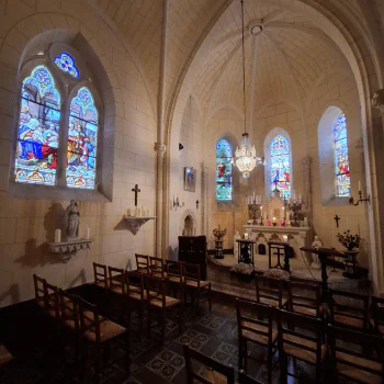 Small chapel interior with wooden chairs, stone walls, a statue, stained glass windows, a chandelier, and an ornate altar decorated with candles and flowers.
